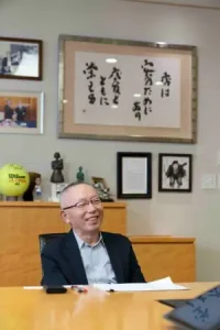 ユニクロを世界企業へ導いたファーストリテイリング柳井正代表取締役会長兼CEO。店舗経営者が学ぶべき商売の原理原則や成功の秘訣について、笑顔でインタビューに応える様子。背後には座右の銘「店は客のためにある」の書。