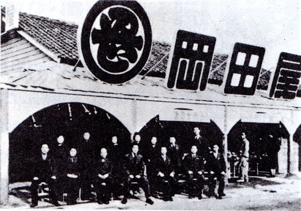 イオンの原点である「岡田屋」創業期の店舗と従業員。倉本長治氏の商人道に基づき、中内功氏らと切磋琢磨した岡田卓也氏の経営哲学の出発点。日本の小売業の歩みと、時代を超えて受け継がれる「商人の志」を伝える貴重な歴史的記録。