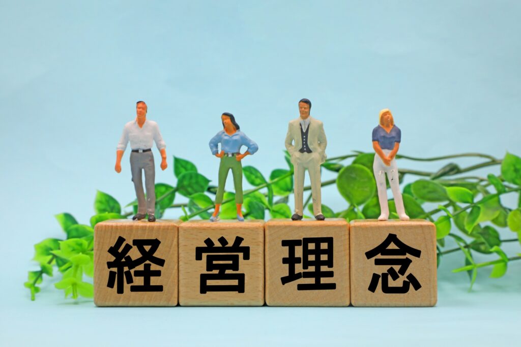 「経営理念」の文字の前に並ぶ人形たち。ドミノ・ピザが学生アルバイトにまで徹底した「30分以内にお届けし、適正な利益を得る」というMission Statementを体現し、組織の方向性を一致させる経営の在り方を示しています。