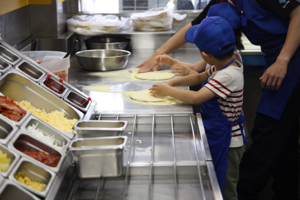 ドミノ・ピザのキッチンで子供たちがピザ生地を伸ばす体験学習の風景。効率的なレイアウトのコールドテーブルとトッピング食材が並ぶ。中内功氏や柳井正氏が重視した「現場での体験」と「標準化された作業環境」が、次世代のファンと担い手を育てる重要性を象徴。