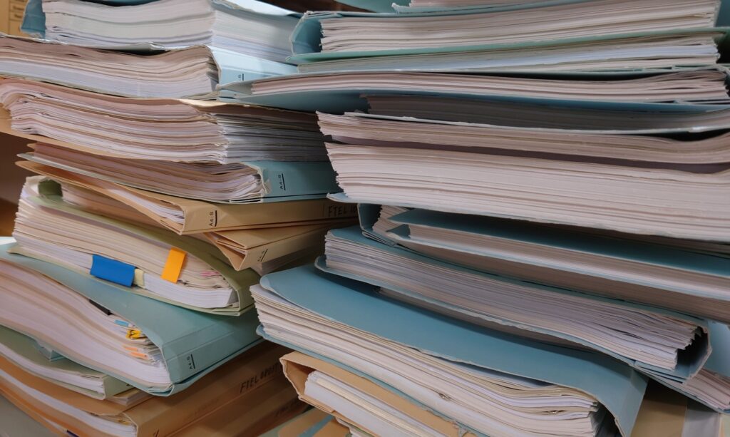 山積みになった大量の帳票ファイルと書類。単なる事務処理に忙殺される現状から脱却し、数字の異常を見極めるマネジメントへの転換を象徴。店長マニュアルで帳票管理を標準化し、経営者視点で業務効率と利益を向上させるための、整理・改善前の現場イメージ