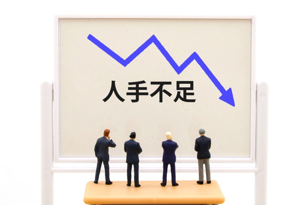 ホワイトボードに描かれた右肩下がりの青いグラフと「人手不足」の文字。深刻な採用難が店舗の運営品質（QSC）を低下させ、業績悪化を招くリスクを警告。監査によってオペレーションの無理・無駄を可視化し、省人化経営への転換を促す。