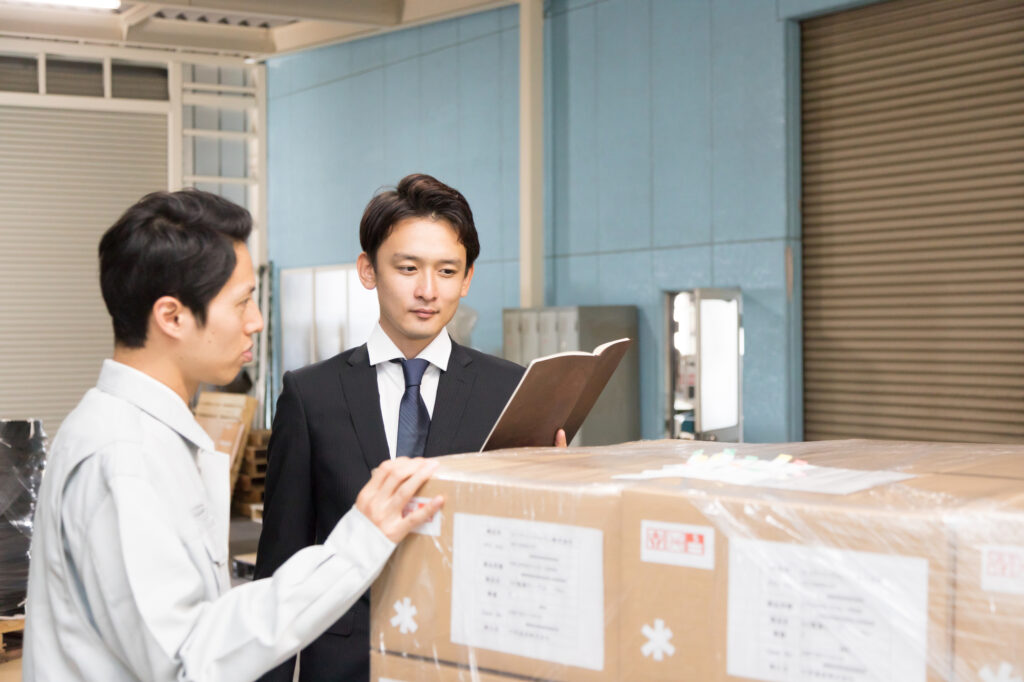 工場や倉庫の現場でスーツ姿の経営者が帳票を確認し、作業スタッフと直接対話する店舗監査の様子。柳井正氏の実践した「現場・現物・現実」の三現主義に基づき、現場の規律とQSCの徹底状況を厳格にチェックする経営ガバナンス。