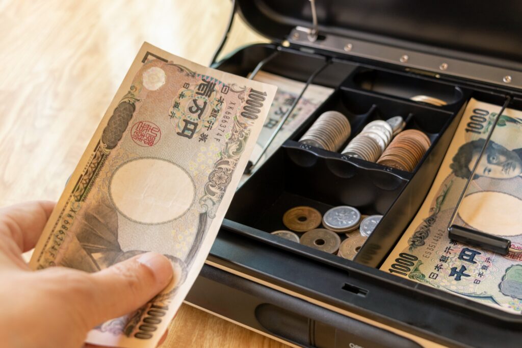 手提げ金庫から取り出した現金と整然と並ぶ硬貨。日々のレジ締め業務を標準化し、1円単位の差異も逃さない正確な帳票管理を強調。ミスを防ぐ前向きなサイクルを回すことで、現場力を高め、最終的な売上アップに繋げるピープル・ビジネスの実践イメージ。