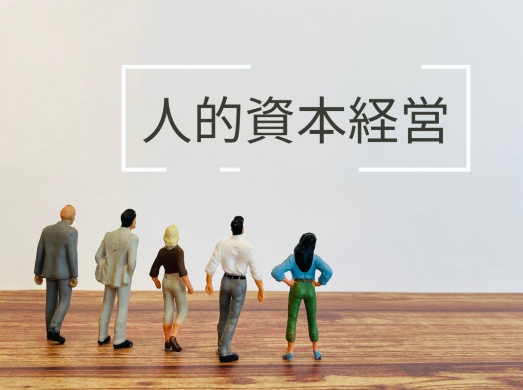 「人的資本経営」の文字を背に、未来を見据えるスタッフのミニチュア。労務管理を単なる事務ではなく、人を「資本」と捉えた投資戦略として実践。スタッフの自発的行動を促し、売上利益の向上と持続的な店舗成長を実現する、ピープル・ビジネスの本質を示す