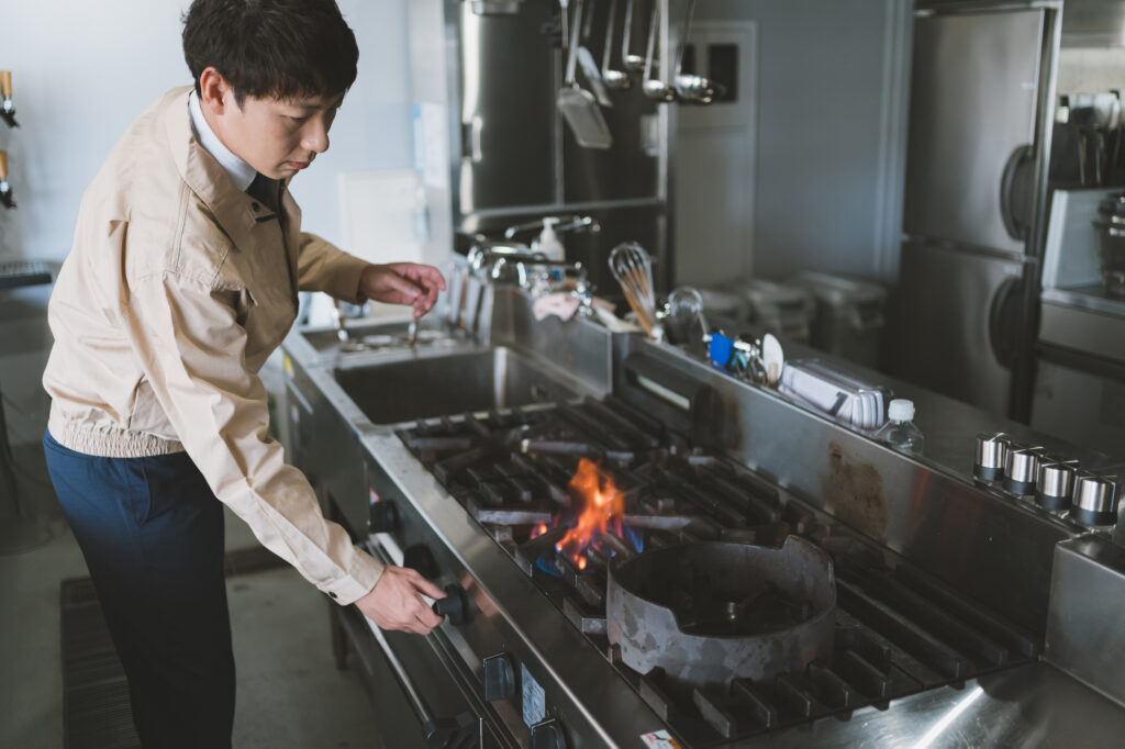 厨房のコンロを点検する店長。店舗経営の根幹である調理インフラの保守は、事故リスクを防ぎ安定稼働を支えます。知識不足による機会損失を防ぎ、効率的な光熱費管理で利益を増やす極意を実践。現場力を高めて「儲かる」店舗をつくるための施設管理の風景。