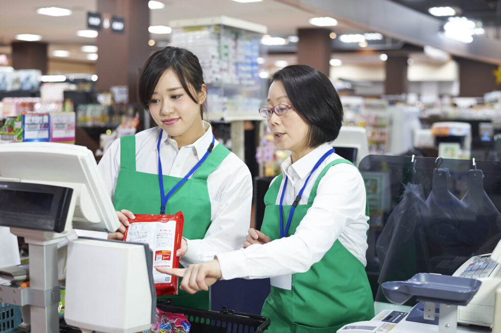 ベテランスタッフが新人にレジ操作を教えるトレーニング風景。教育訓練の基礎を標準化し、知識不足によるトラブルを未然に防ぎます。理念を掲げた丁寧な接客スキルを伝承し、人間力を磨くことで前向きな店舗運営サイクルを回す、人材育成の基本イメージ。