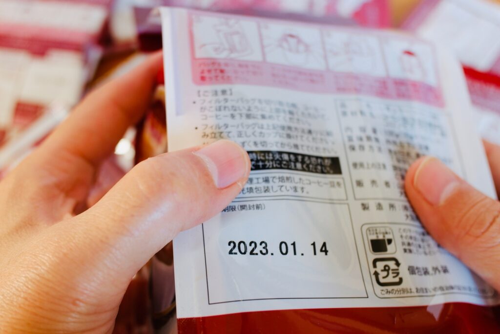 商品の背面に印字された消費期限「2023.01.14」を指で指し、厳格に確認している様子。食品衛生法に基づく顧客との約束である期限表示と、シールの貼り替えや二重貼りといった安易な偽装が経営を破滅させるリスクを想起させる。