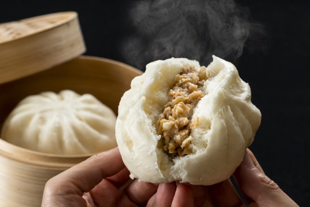 蒸し器から取り出された、湯気が立ち上る熱々の肉まん。中身の具材が豊かにはみ出し、食欲をそそるシズル感を強調。中内功や岡田卓也が重視した「季節の先取り」と「五感に訴える販売」を象徴する、店舗経営の主力商品のイメージ。