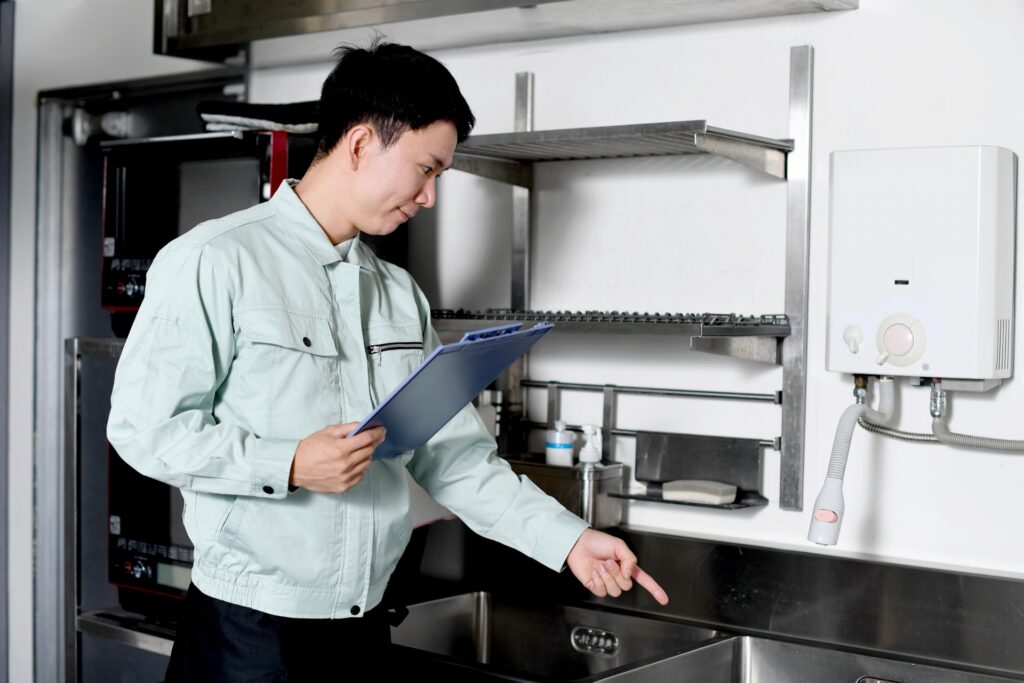 作業着の男性が厨房でクリップボードを手に、シンクや手洗設備を指差し確認している様子。ミニストップの不祥事を教訓に、本部のSVや品質保証部が徹底すべき厳格な店舗監査と、不正を未然に防ぐ現場点検の重要性を象徴するイラストACの素材。
