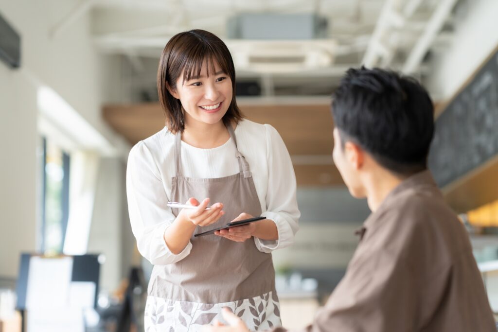 カフェで笑顔で注文を取るスタッフと、寄り添うリーダー。人財力を活かした「売上を上げられる人の育成」により、組織全体の接客レベルを底上げ。戦略的な声掛けや提案により客単価をアップさせ、パート・アルバイトと一丸となって繁盛店を築く実践風景。