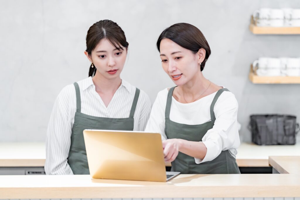カフェのカウンターでPC画面を見ながらスタッフと業務改善を話し合う店長。教育訓練を標準化し、実務と評価を連動させることで人財力を最大化。知識不足による不安を取り除き、前向きな育成サイクルで繁盛店を築くための実践的なトレーニングイメージ。