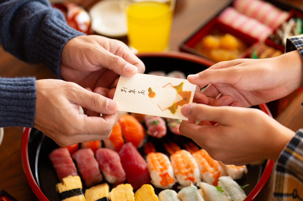 お正月に家族で寿司を囲み、子供にお年玉を手渡す温かい光景。2026年の正月休業を象徴する、従業員が家族との時間を大切にする「働き方の質」の向上をイメージしたPixtaの素材。