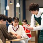 店舗でのOJT（現場研修）シーン。店長候補の新入社員が実務習得を通じてパート・アルバイトと信頼関係を築く様子。頭でっかちな思考を捨て、謙虚に現場を学ぶ姿勢が店全体のコミュニケーションとマネジメント能力向上に繋がるプロセスを表現した画像。