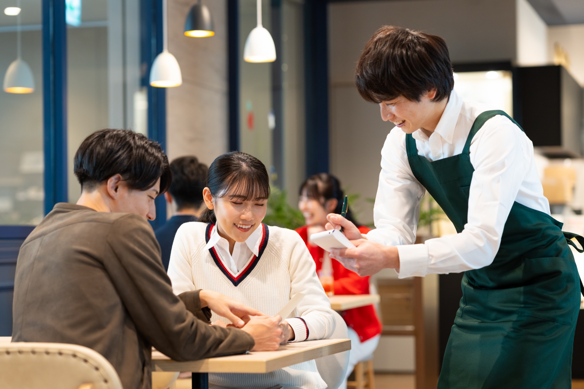 店舗でのOJT(現場研修)シーン。店長候補の新入社員が実務習得を通じてパート・アルバイトと信頼関係を築く様子。頭でっかちな思考を捨て、謙虚に現場を学ぶ姿勢が店全体のコミュニケーションとマネジメント能力向上に繋がるプロセスを表現した画像。