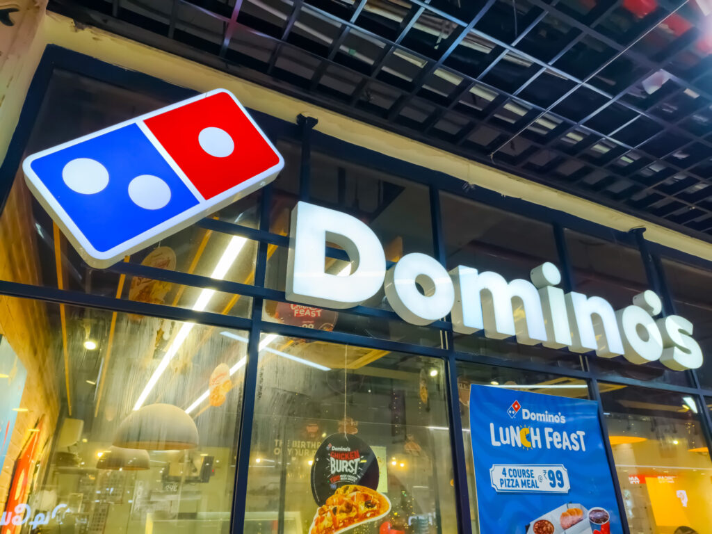 「Domino's」が輝くドミノ・ピザの店舗看板。米創業者トム・モナハンの哲学「完璧なピザを30分以内に届ける」に基づき、繁忙期の夜間でも品質と配送時間を守るためのキャパシティマネジメントと、SNS炎上を回避するオペレーション戦略の象徴。