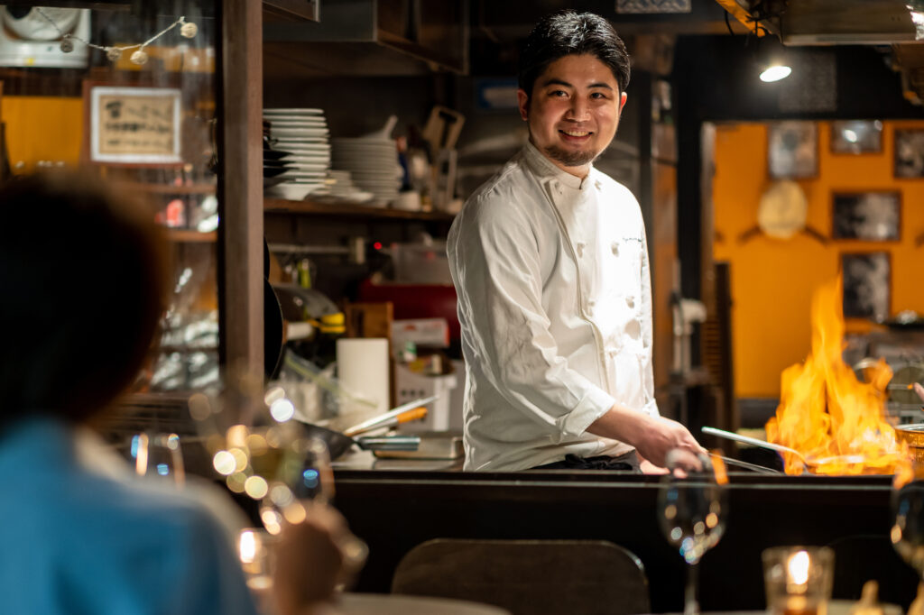 厨房で調理に集中するプロのシェフ。個人店が人手不足対策を完遂するために、無駄な動きを削ぎ落としたコックピット型の動線設計を構築し、一歩も動かずに主要な調理を完結させる「足数」削減による圧倒的な効率化と、プロの技術の融合を表現。