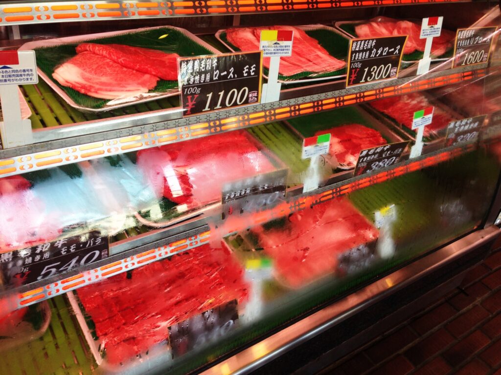 精肉店の冷蔵・冷凍ショーケース。夏の猛暑期における突発的な停電や、それに伴う食材廃棄ロス、太陽光発電や蓄電池による優先給電設計の必要性を視覚的に伝える精肉売場の写真。