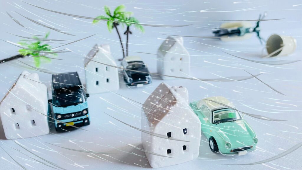 強風や暴風雨にさらされる家と車のミニチュア。台風シーズンである7月に、店舗が直面する浸水やのぼり旗・看板の飛散などの気象災害リスクを想起させ、店舗防衛を促すイメージ写真。
