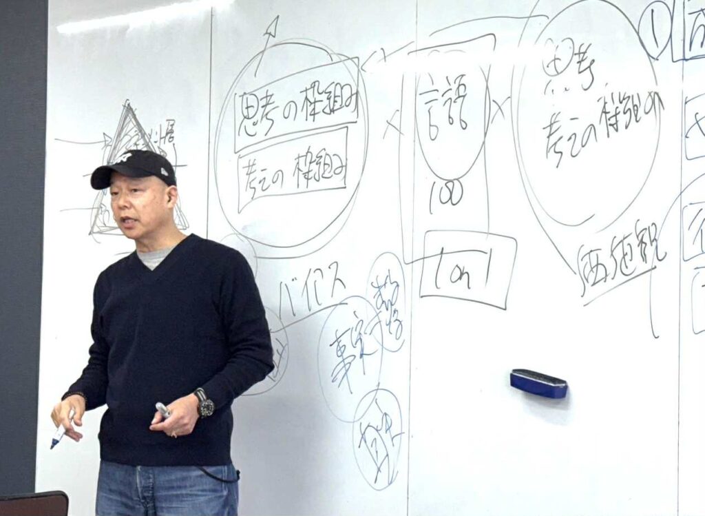 ホワイトボードの前で自走サイクルを解説する佐藤勝人。視点や思考の枠組みを変え、新しい道具やITシステムを導入することで自動的に現場の行動と意識を変革するプロセスを論じる。
