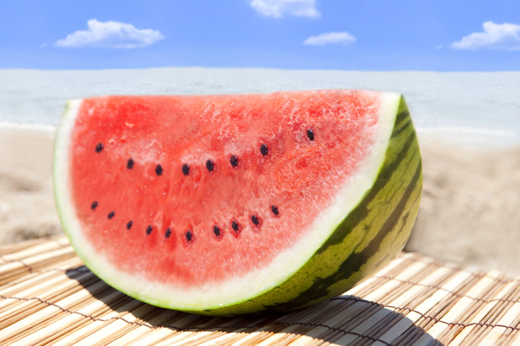 7月の小売店・飲食店における夏商戦の売り場作りや、サマーセール・クリアランスのイメージを表現する、夏の海とカットされたみずみずしいスイカのディスプレイ写真。