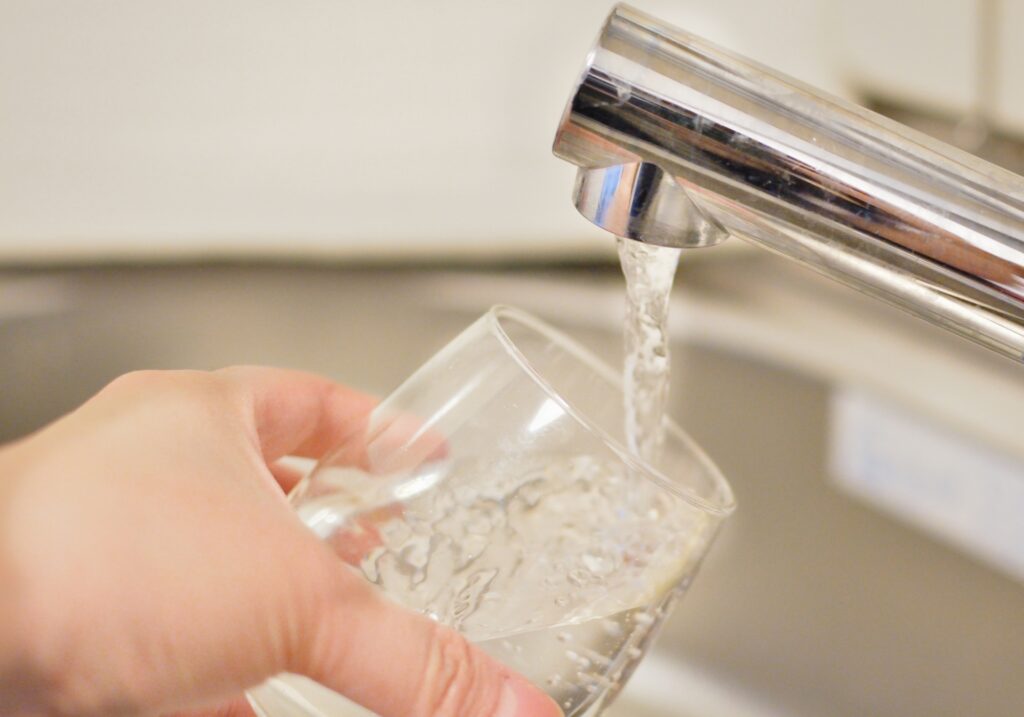 水道蛇口からグラスに注がれる水。老朽化インフラの破損に伴う大規模断水に備え、店舗の衛生管理・生活用水の備蓄や、使い捨て食器等を活用したドライ運用のBCP(事業継続計画)手順を提示。