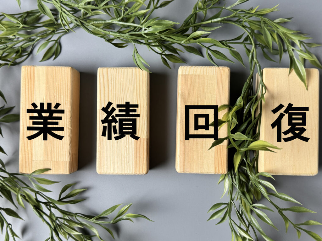 緑の植物に囲まれた、木製ブロックに書かれた「業績回復」の文字。物価高やコスト高騰の荒波を乗り越え、経営理念を掲げて日々の実務と連動させ、評価する前向きなサイクルを社内に回すことで強固な売上基盤を完成させるイメージ。