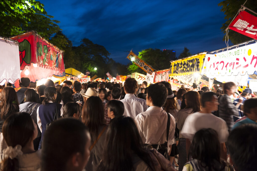 夏の集客イベントやお祭りで大混雑する店舗周辺の人出と屋台のイメージ。突然の将棋倒しや事故を防ぐため、事前の避難動線確保と人員配置、安全管理の徹底で店舗と顧客を守るリスク管理の実践手順。