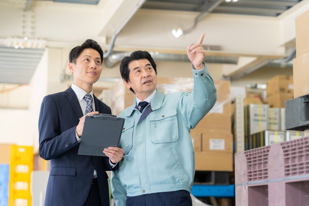倉庫内で荷物を指差しながら打ち合わせを行う経営者と取引先パートナー。単なるコスト削減の下請け関係を超え、有事に優先供給や物資融通を可能にする「取引先は財産」という理念に基づく共存共栄の協力体制を表現した風景。