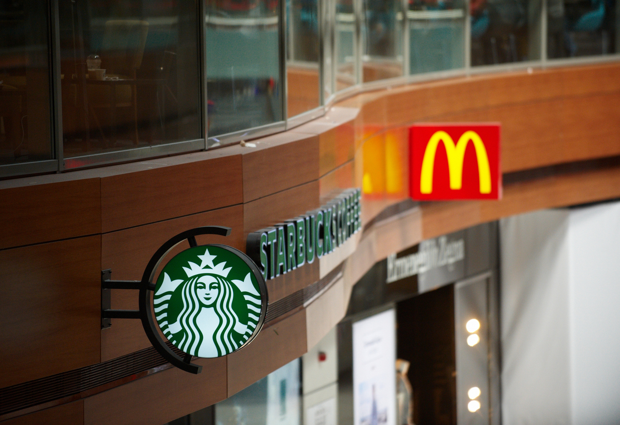 世界的な飲食チェーンであるスターバックスとマクドナルドの店舗看板。創業期の主力商品名表記からロゴ単体へとシンプル化を進めたメガブランドの戦略的背景と、藤田田氏が実践した業態認知の重要性を象徴する店頭ファサード。