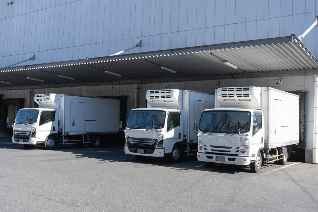 物流倉庫の搬入口に並ぶ配送トラック。自店舗に直接の被害がなくても、仕入先や流通拠点の被災によって商品供給が止まるサプライチェーン途絶リスクを示し、代替仕入れルート確保と複数購買の必要性を伝える実務イメージ。
