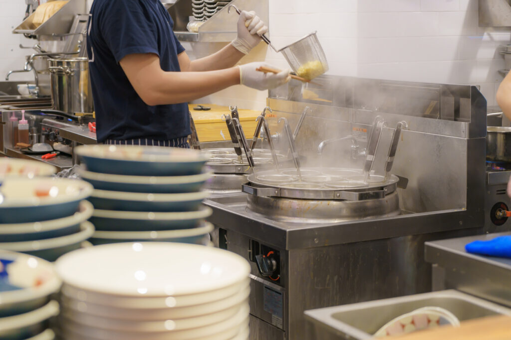 大量の丼が積まれ湯切り作業に追われる飲食店の厨房。直前の駆け込みや当日フリー客に頼る店舗運営は、調理遅延や食材廃棄、突発的な人件費高騰を招き、粗利益を著しく圧迫するため事前予約による平準化が不可欠となる。