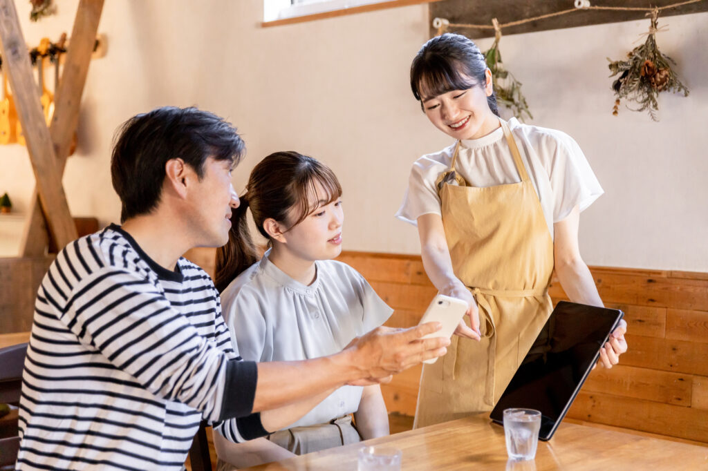 飲食店でスタッフがタブレット端末を用いて来店客のスマホ注文を案内する接客風景。日本マクドナルド創業者・藤田田氏の作業動線効率化に倣い、ピーク時の混乱防止と客単価向上を両立する実務オペレーション。