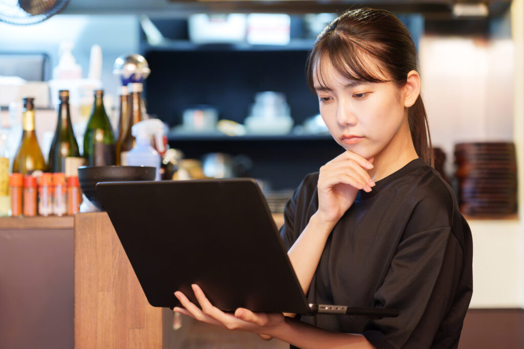 仮説検証の原則に基づき、店舗の売上データや日々の営業実績をパソコン画面で冷静に分析・検証する女性店長