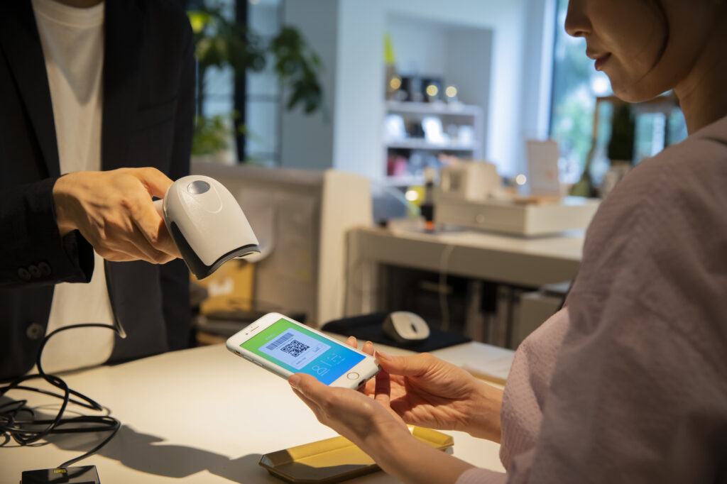 店頭レジでスマートフォンのバーコード画面を読み取るキャッシュレス・クーポン受付風景。店頭卓上POPとLINE公式アカウントを連動させ、人手不足の現場でも会計動線を標準化して回転率を高める仕組み。
